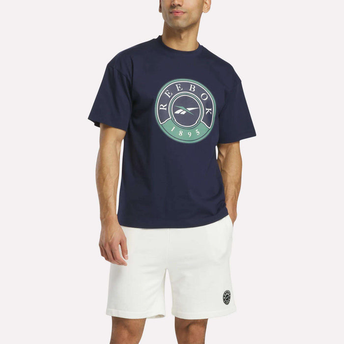 A man stands against a plain light background wearing a navy blue Reebok Men's Lawn Club T-Shirt with a circular logo and white shorts.