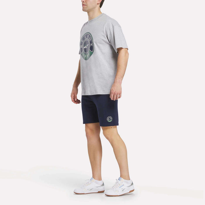 A man stands wearing a light grey Reebok Men's Lawn Club T-Shirt, navy blue shorts with a small emblem, and white athletic shoes against a plain white background.