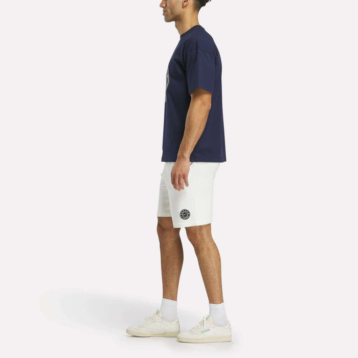 A man stands in profile wearing the Reebok Men's Lawn Club T-Shirt, white shorts with a Lawn Club logo, white socks, and white sneakers against a plain light background.