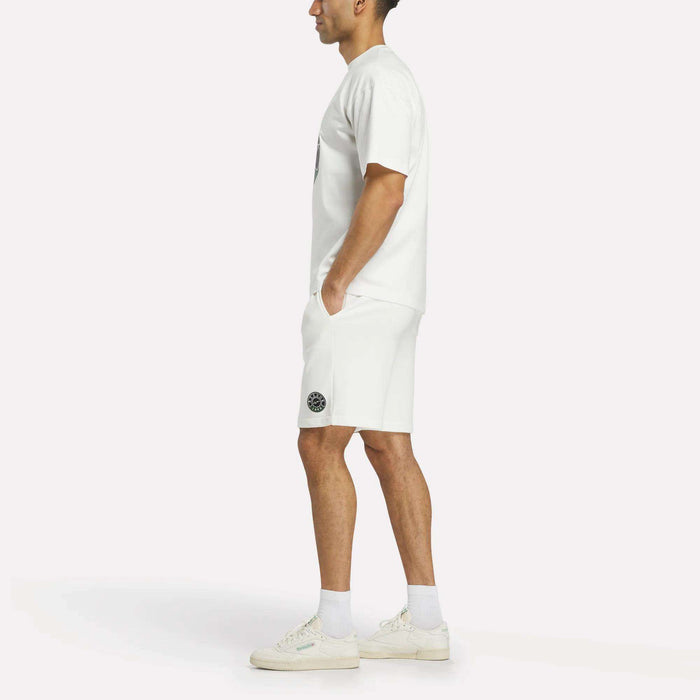 A man stands in profile wearing the Reebok Men's Lawn Club T-Shirt, white shorts with a black patch on the left leg, white socks, and white sneakers against a plain light background—ideal for the Lawn Club.