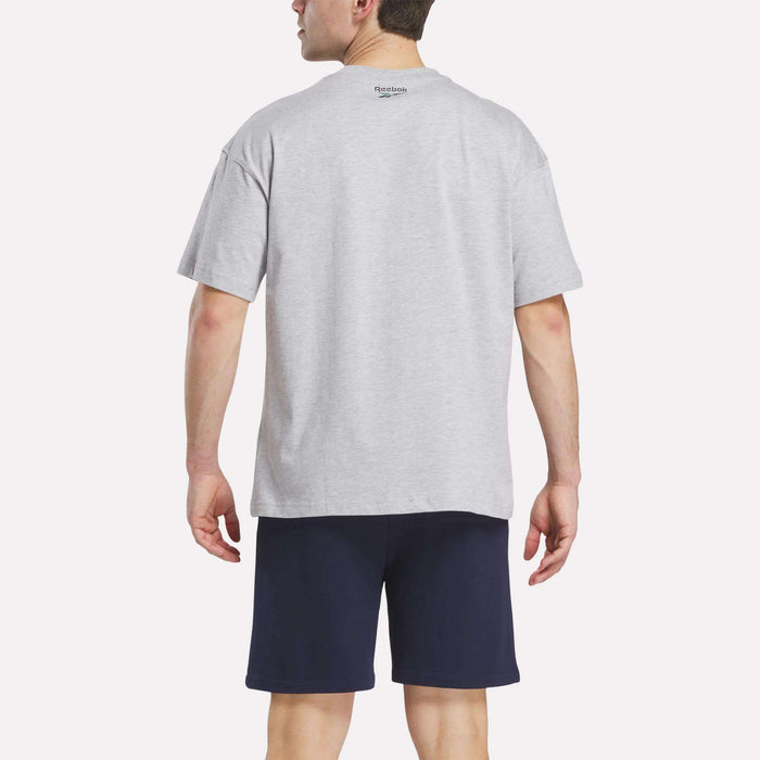 A man is seen from the back wearing a light gray Reebok Men's Lawn Club T-Shirt and navy blue shorts against a plain, light background.