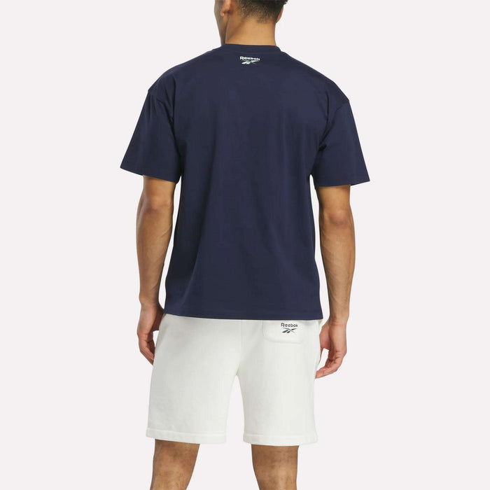 A person faces away, wearing a navy blue Reebok Men's Lawn Club T-Shirt made of luxe cotton and white shorts. The Lawn Club logo is on the back of the shirt near the neck and on the waistband of the shorts.