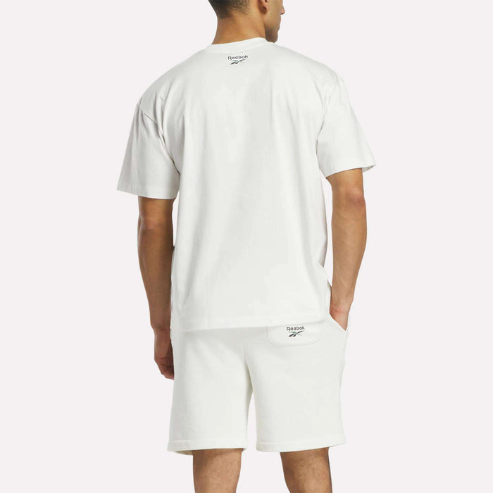 A man faces away in a loose-fit Reebok Men’s Lawn Club T-Shirt and white shorts, both featuring small Reebok logos on the back, set against a plain light background.