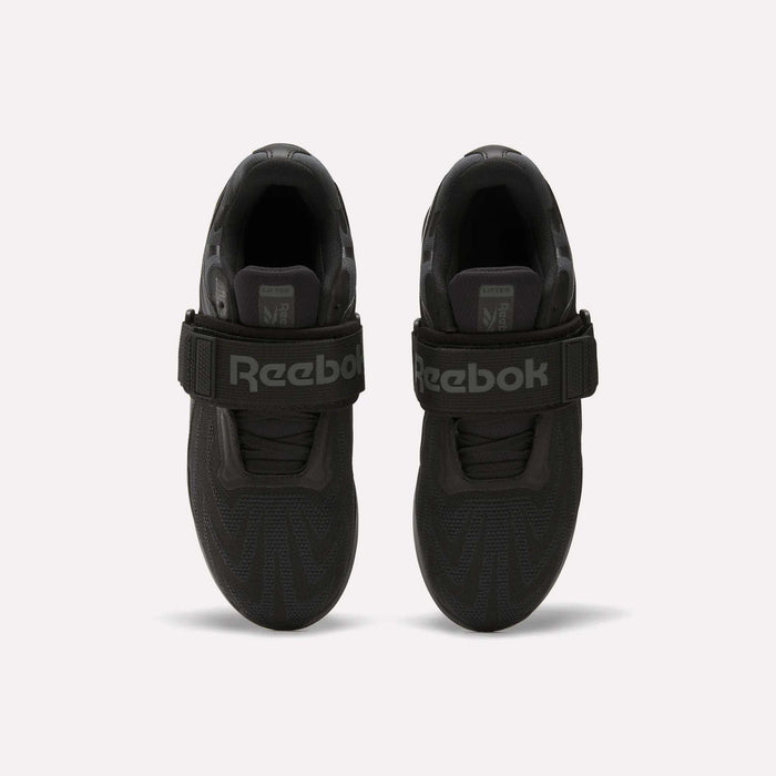 Top view of a pair of Reebok Men's Legacy Lifter IV Shoes in black, with thick textured soles and wide velcro straps for stability. The large Reebok logo is visible. Set against a plain, light-colored background.