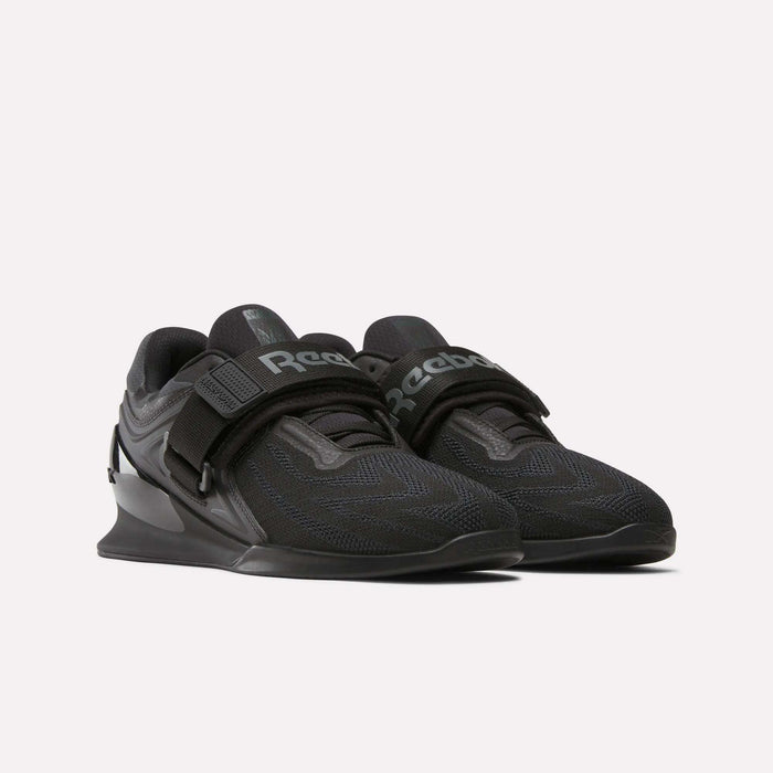The Reebok Men's Legacy Lifter IV Shoes feature a thick wedge heel, mesh upper, and wide midfoot strap for enhanced stability. Reebok branding is visible on the tongue and strap, shown against a plain white background.