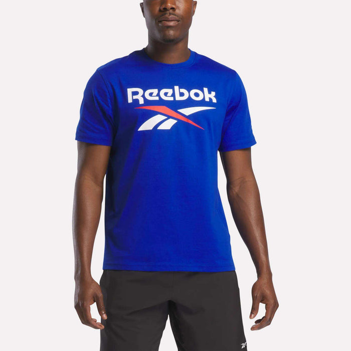 A person wearing the Reebok Men's Logo Crewneck T-Shirt in bright blue, featuring a large white and red vector logo across the chest, paired with black shorts, stands against a plain white background.