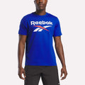 A person wearing the Reebok Men's Logo Crewneck T-Shirt in bright blue, featuring a large white and red vector logo across the chest, paired with black shorts, stands against a plain white background.