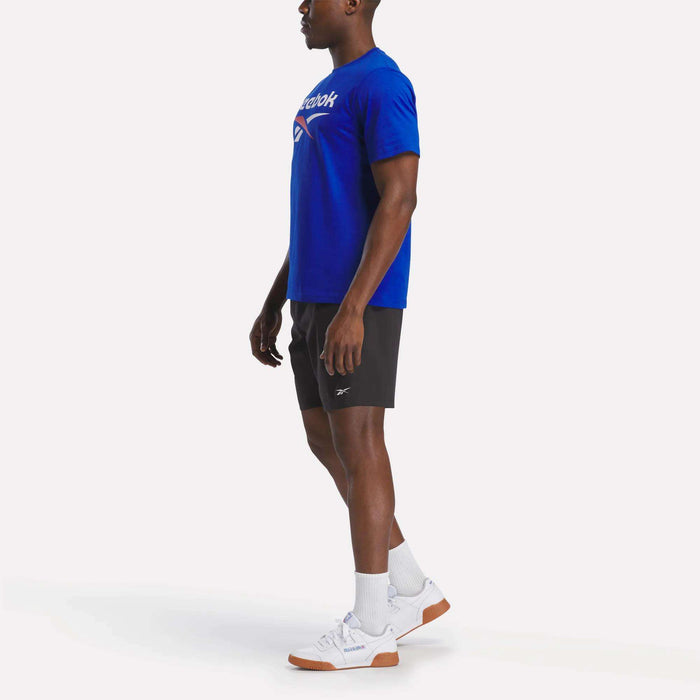 A man faces left in a blue Reebok Men's Logo Crewneck T-Shirt, black shorts, white crew socks, and white athletic shoes with brown soles, standing against a plain white background.