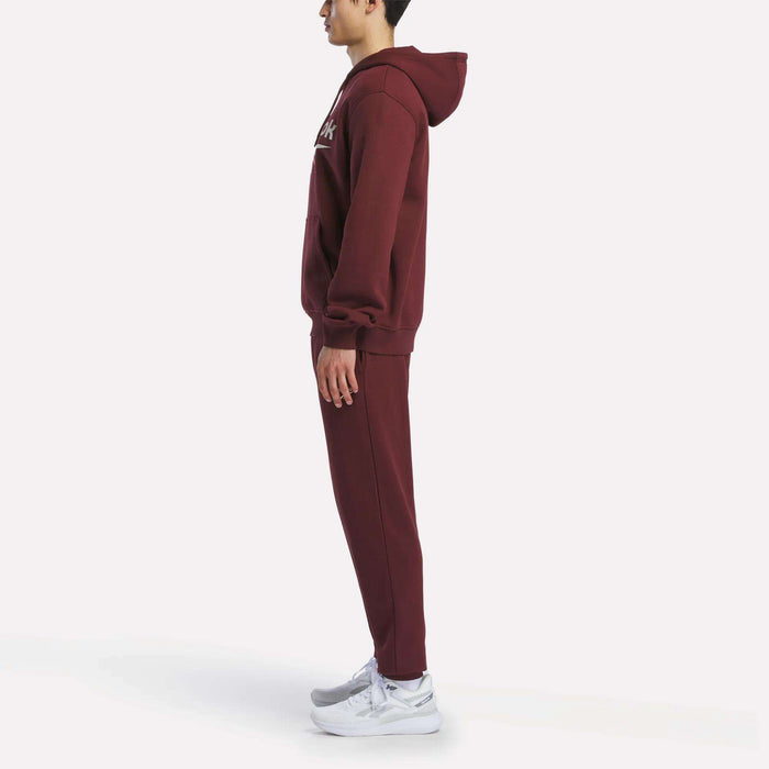 A man in profile wears a maroon Reebok Men's Logo Hoodie and matching sweatpants with white sneakers, standing against a plain white background.