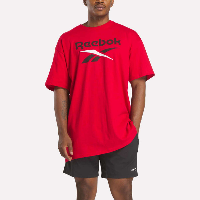A person wears a loose red Reebok Men's Stacked Vector T-Shirt with a bold logo, paired with black athletic shorts featuring a small Reebok logo, standing against a plain white background.