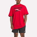 A person wears a loose red Reebok Men's Stacked Vector T-Shirt with a bold logo, paired with black athletic shorts featuring a small Reebok logo, standing against a plain white background.