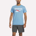 A man stands facing forward against a plain white background, wearing a light blue heather Reebok Men's Stacked Vector Graphic T-Shirt and dark gray athletic shorts.