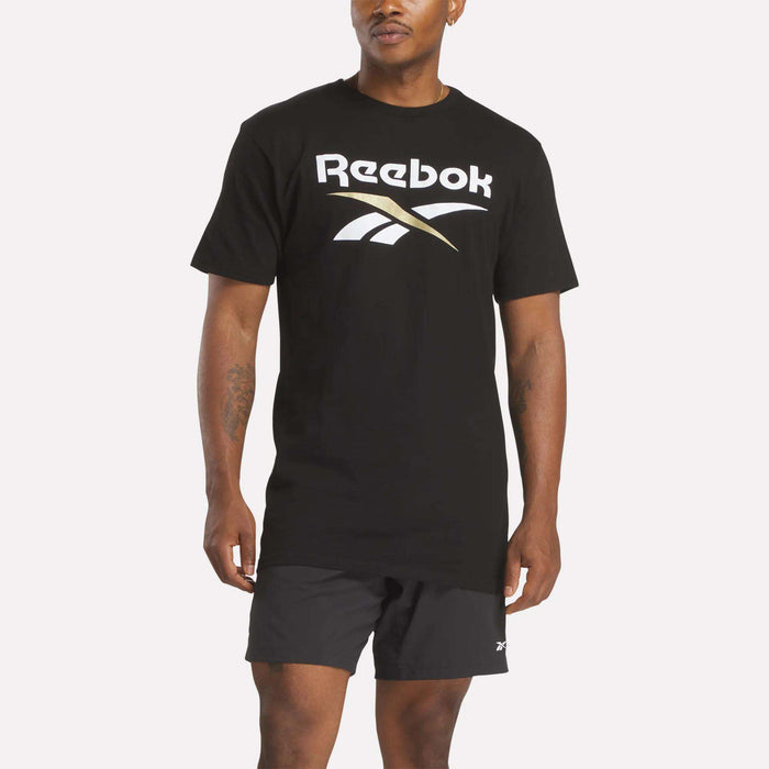 A person models the Reebok Men's Stacked Vector T-Shirt in black, featuring a bold white and gold logo, paired with matching black shorts, set against a plain white background.