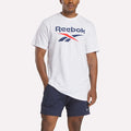 A man wears a white Reebok Men's Stacked Vector T-Shirt featuring a large logo on the chest and navy blue shorts, standing with one hand in his pocket against a plain light background.