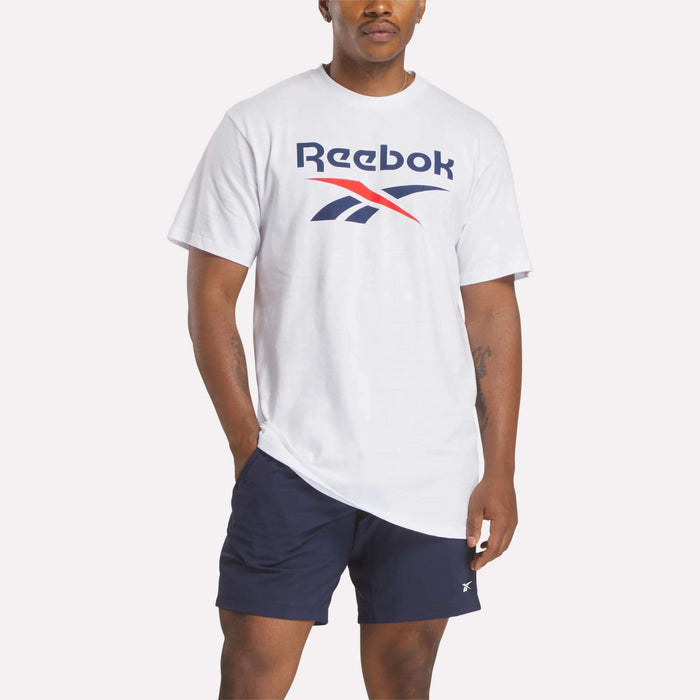 A man wears a white Reebok Men's Stacked Vector T-Shirt featuring a large logo on the chest and navy blue shorts, standing with one hand in his pocket against a plain light background.