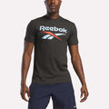 A man in a dark gray Reebok Men's Stacked Vector Graphic T-Shirt, 100% cotton with a bold white and red chest logo, paired with navy blue shorts, stands against a plain light background.