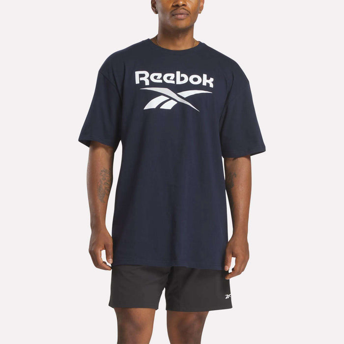 A man wears a loose-fitting Reebok Men's Stacked Vector T-Shirt in navy blue cotton with a bold white logo, paired with black athletic shorts, standing against a plain white background.