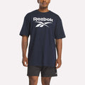 A man wears a loose-fitting Reebok Men's Stacked Vector T-Shirt in navy blue cotton with a bold white logo, paired with black athletic shorts, standing against a plain white background.