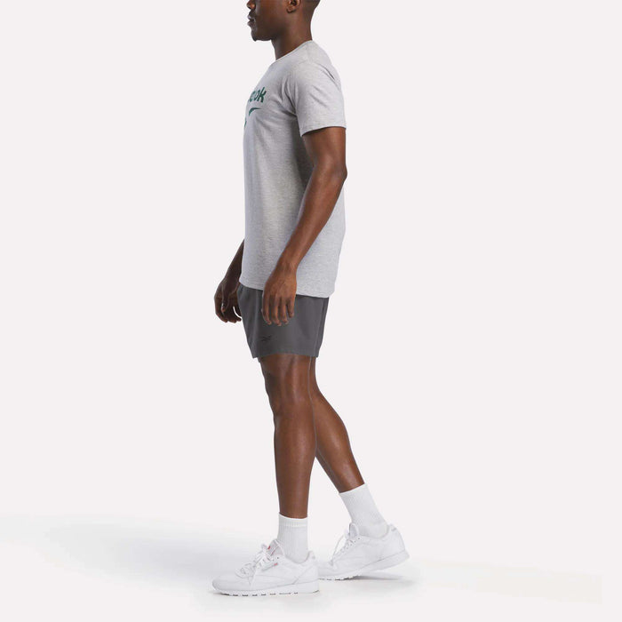 A man stands in profile against a plain background, wearing a gray Reebok Men's Stacked Vector Graphic T-Shirt, dark gray shorts, white socks, and white athletic shoes.