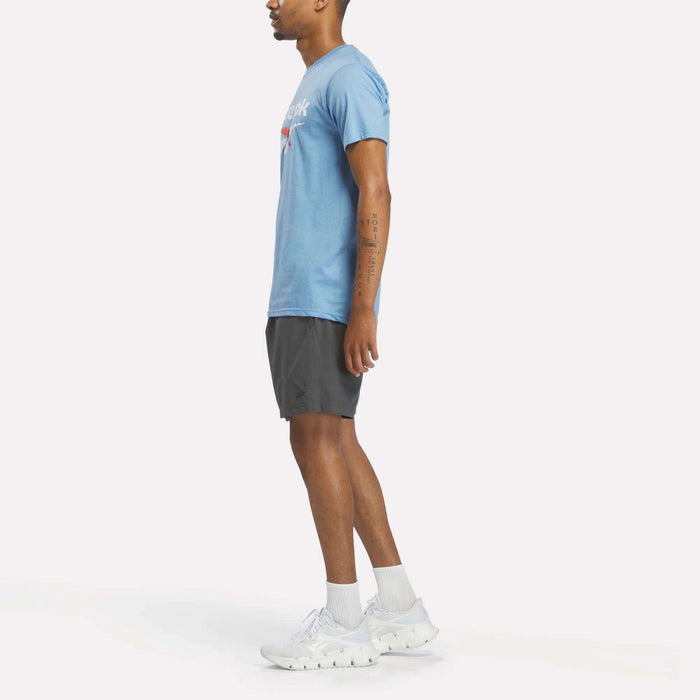 A person stands in profile facing left, wearing a light blue Reebok Men's Stacked Vector Graphic T-Shirt, dark athletic shorts, white socks, and white Reebok sneakers against a plain white background.