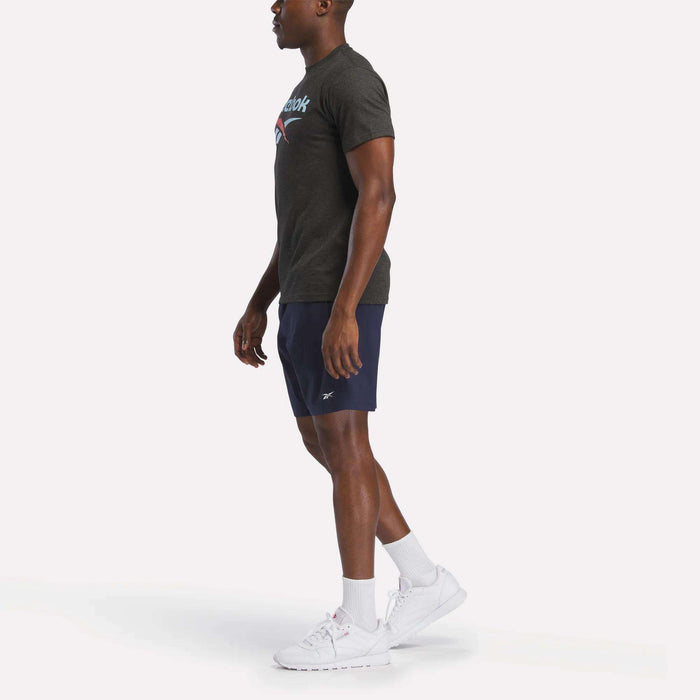 A man in profile wears a lightweight, dark grey Reebok Men's Stacked Vector Graphic T-Shirt, navy shorts, white crew socks, and white sneakers against a plain white background.