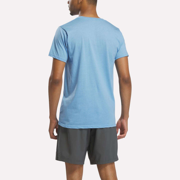 A person stands with their back to the camera, wearing a Reebok Men's Stacked Vector Graphic T-Shirt in light blue and dark gray shorts, set against a plain light background.