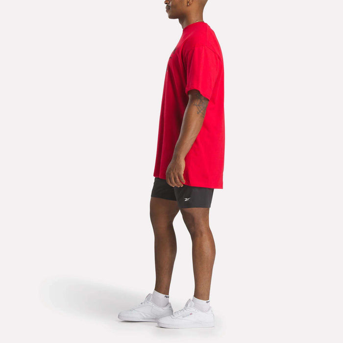 A man stands in profile against a white background, wearing a loose red Reebok Men's Stacked Vector T-Shirt, black shorts, and white sneakers.