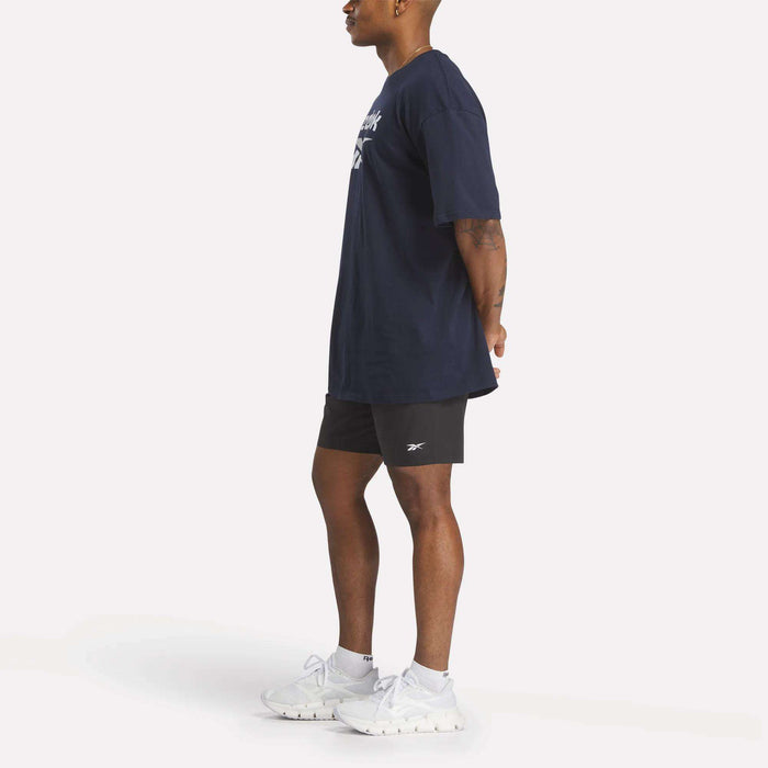 A person stands in profile with hands behind their back, wearing a navy Reebok Men's Stacked Vector T-Shirt, black shorts, white socks, and white Reebok athletic shoes against a plain white background.