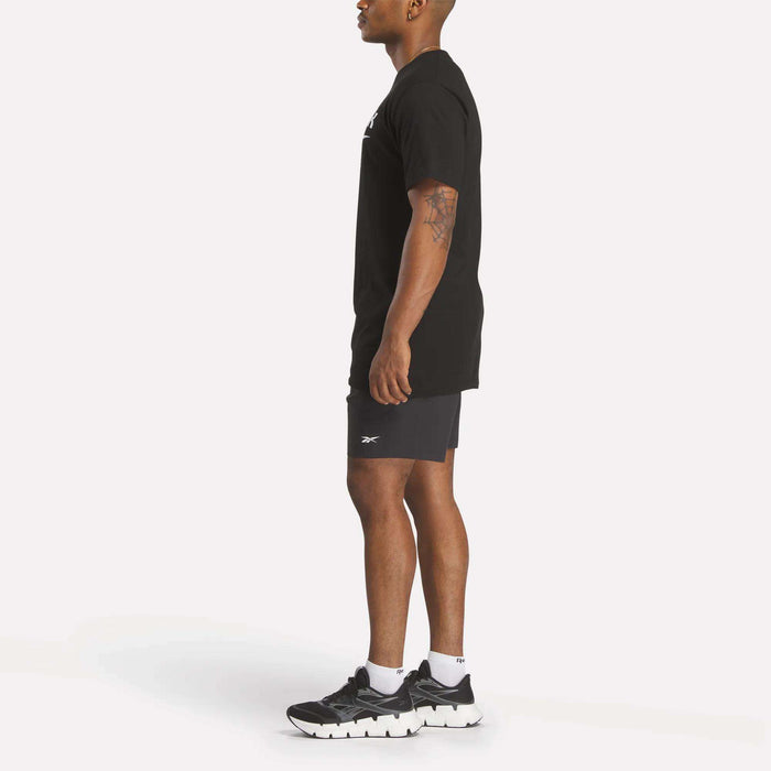 A man stands in profile against a plain background wearing a black Reebok Men's Stacked Vector T-Shirt, black shorts, white socks, and black athletic shoes. His arms are relaxed at his sides with a tattoo visible on his left arm.