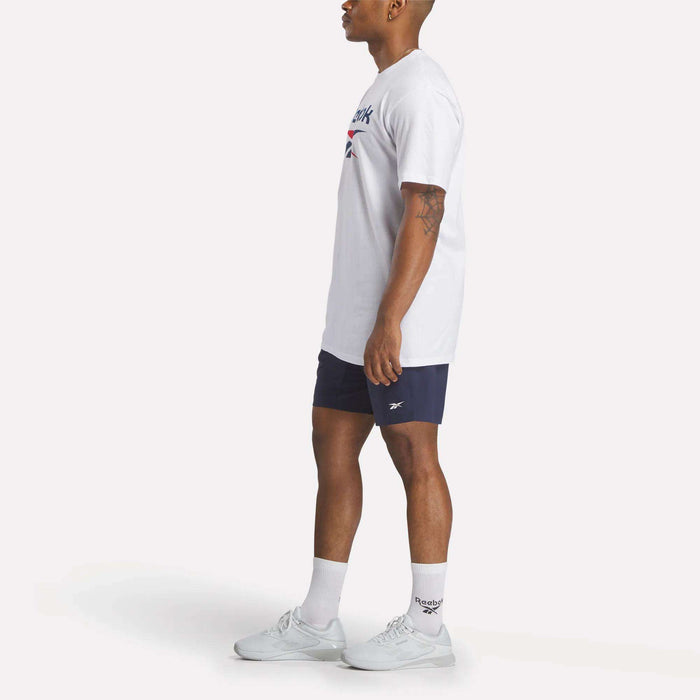A man stands in profile against a plain light background wearing a Reebok Men's Stacked Vector T-Shirt, navy Reebok shorts, white crew socks, and white sneakers.