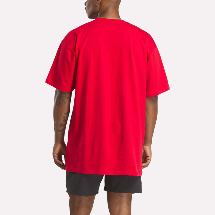 A person faces away from the camera, wearing a Reebok Men's Stacked Vector T-Shirt in bright red and black shorts. The background is plain white.
