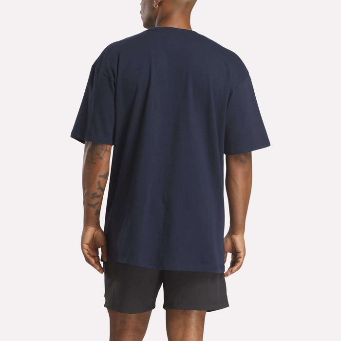 A person stands with their back to the camera, wearing a loose-fit Reebok Men's Stacked Vector T-Shirt in navy blue and black shorts, showing short hair and tattoos on their left arm against a plain white background.