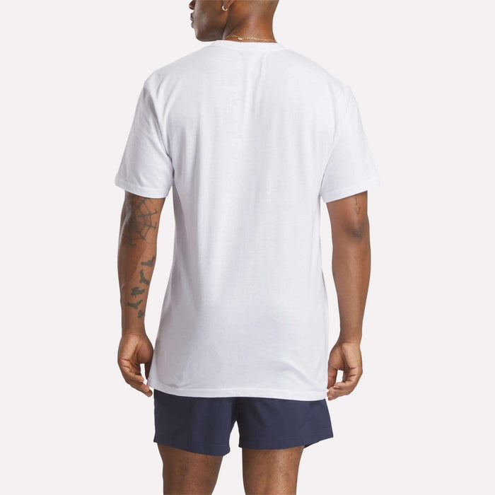 A person with a visible arm tattoo stands facing away, wearing the Reebok Men's Stacked Vector T-Shirt in plain white and navy blue shorts, against a simple light-colored background.
