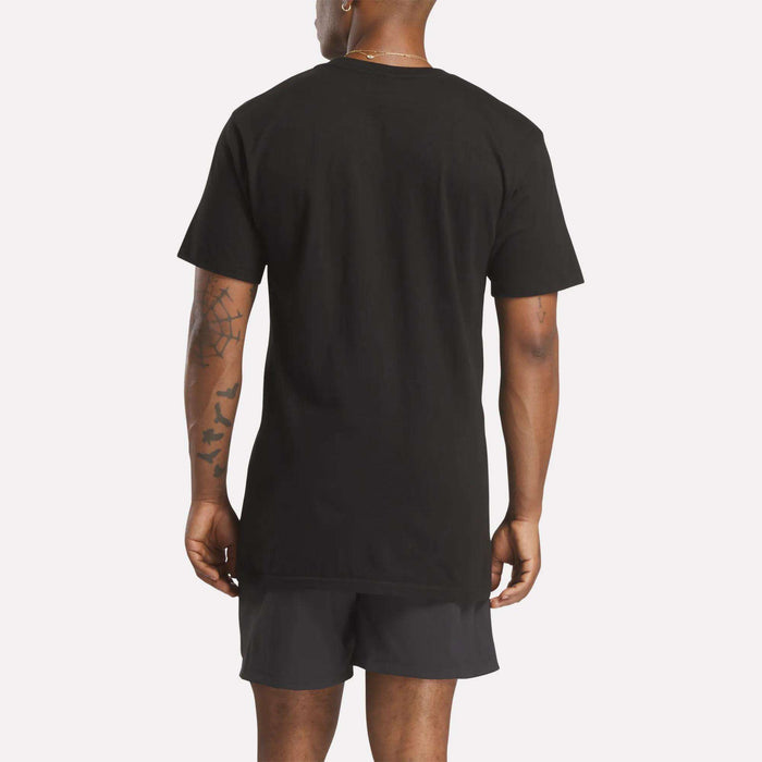 A person stands with their back to the camera, wearing a plain black Reebok Men's Stacked Vector T-Shirt and black shorts. Tattoos are visible on their left arm and both forearms. The background is a solid light color.