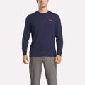 A man wears a Reebok Men's Long Sleeve Golf Sweatshirt in navy blue with UPF 30+ and a small red and white chest logo, paired with light gray pants, standing against a white background.