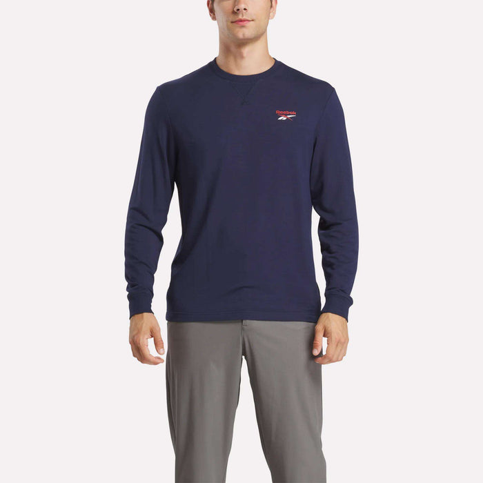 A man wears a Reebok Men's Long Sleeve Golf Sweatshirt in navy blue with UPF 30+ and a small red and white chest logo, paired with light gray pants, standing against a white background.