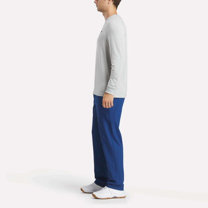 A man in profile wears a Reebok Men's Long Sleeve Golf Sweatshirt in light gray with UPF 30+, blue pants, and white sneakers, standing against a plain white background.
