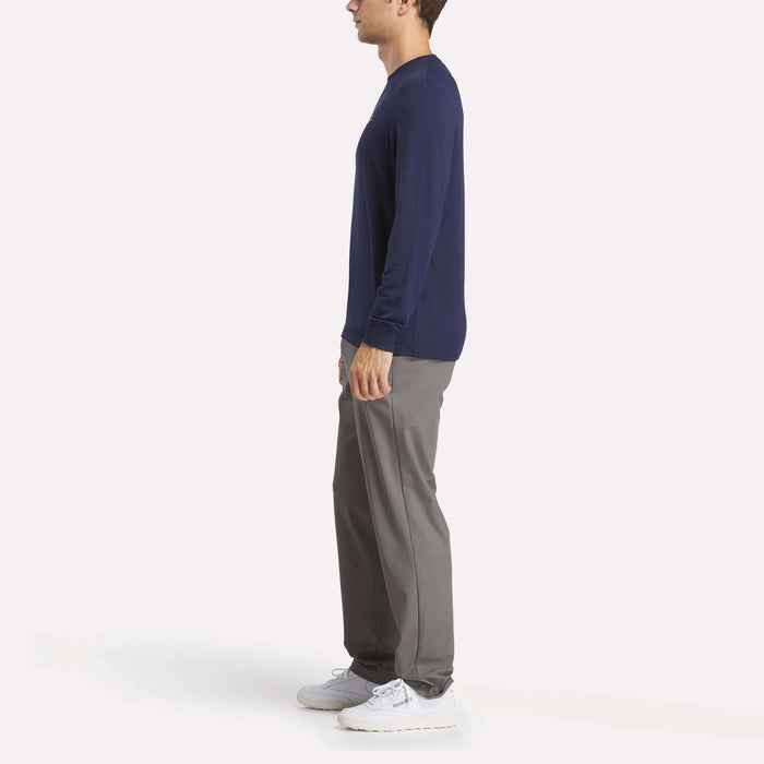 A man in profile stands against a plain white background wearing a Reebok Men's Long Sleeve Golf Sweatshirt in navy, gray pants, and white sneakers.
