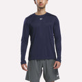 A person wearing a Reebok Men’s Long Sleeve Tech T-Shirt in Vector Navy and gray Reebok shorts stands facing forward against a plain white background.