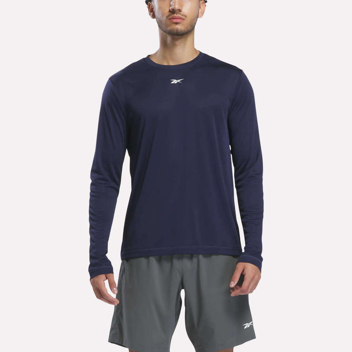 A person wearing a Reebok Men’s Long Sleeve Tech T-Shirt in Vector Navy and gray Reebok shorts stands facing forward against a plain white background.