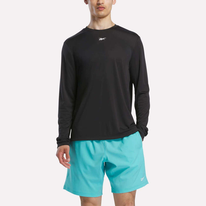 A person wearing a black Reebok Men's Long Sleeve Tech T-Shirt and light blue shorts stands against a plain white background, ready for cold weather workouts.