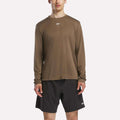 A man in a Reebok Men's Long Sleeve Tech T-Shirt (brown) and black shorts faces forward against a plain light background. Both cold weather workout pieces feature small white logos on the chest and leg.
