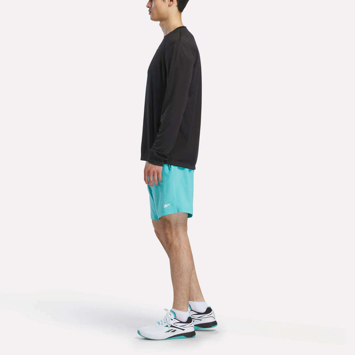 A man stands sideways against a white background, wearing a black Reebok Men's Long Sleeve Tech T-Shirt, turquoise athletic shorts, and white sneakers with black and teal accents—ideal for cold weather workouts.
