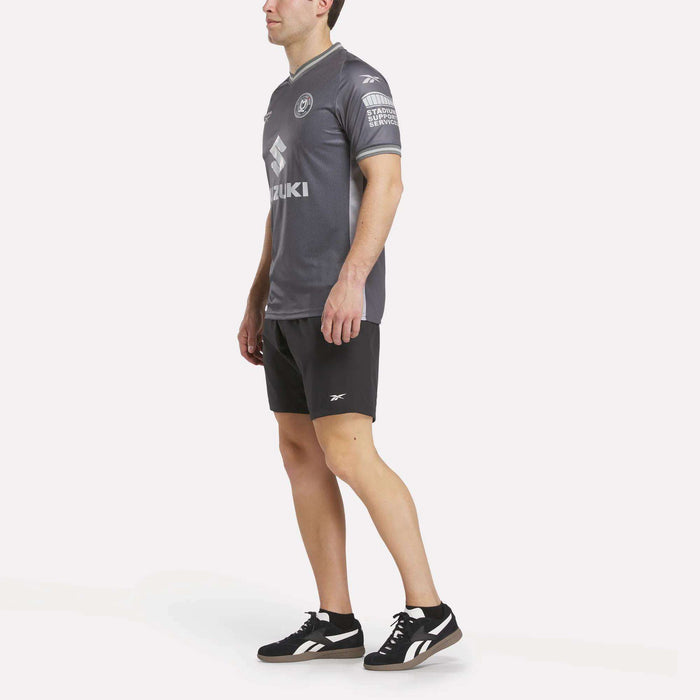 A man wearing a Men's MK Dons Away Jersey, black Reebok shorts, and black Reebok sneakers stands against a plain white background, facing slightly to the side with his arms relaxed by his sides.