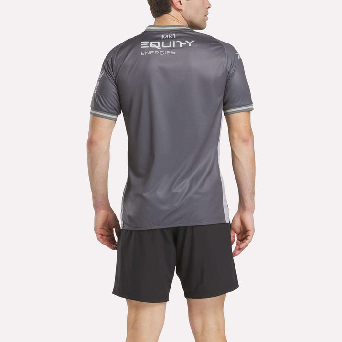 A man faces away, wearing a gray Reebok Men's MK Dons Away Jersey with "MK1 EQUITY ENERGIES" on the back and black athletic shorts, set against a plain light background.