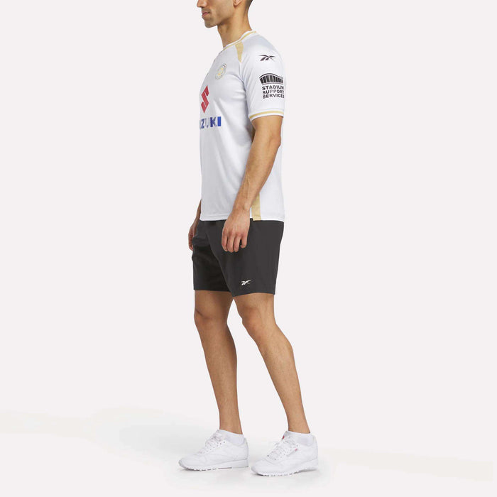 A man stands sideways against a white background wearing the Reebok Men's MK Dons Home Jersey, paired with black shorts and white sneakers.