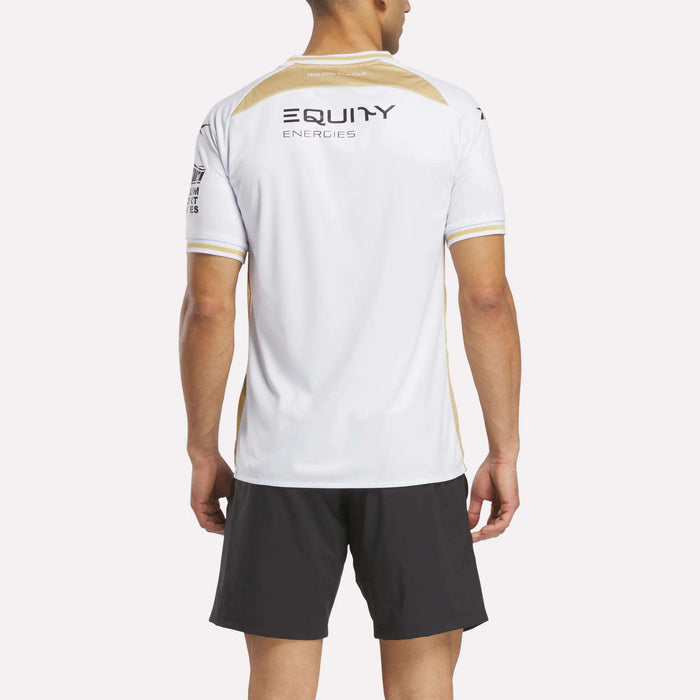A person faces away, wearing a white Reebok Men's MK Dons Home Jersey with EQUITY ENERGIES on the back and black shorts. A plain, light background reflects the vibrant spirit of Milton Keynes football.