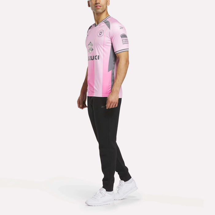 A man stands slightly sideways against a white background, wearing the Men's MK Dons Third Jersey by Reebok in pink and gray, paired with black pants and white sneakers.
