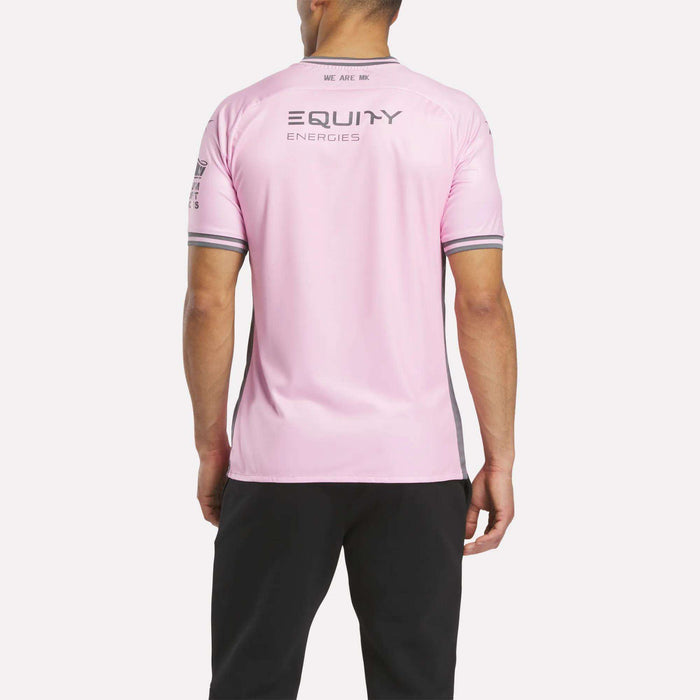 A person in Milton Keynes faces away, wearing a Reebok Men's MK Dons Third Jersey in light pink with gray trim and "EQUITY ENERGIES" on the back, paired with black pants.