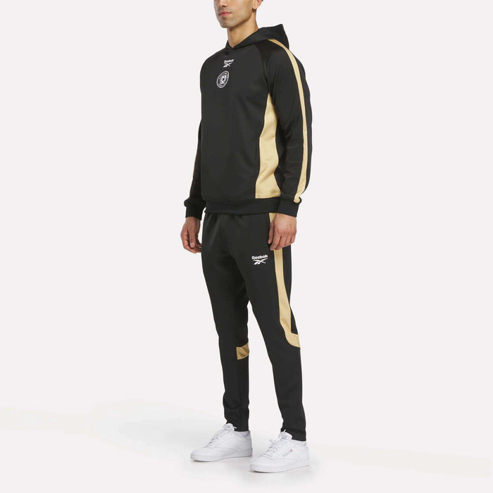 A man wears the Reebok Men's MK Dons Travel Hoodie tracksuit in black with gold stripes on the sleeves and legs, a hood, and a white Reebok logo, paired with white sneakers. The background is plain white.